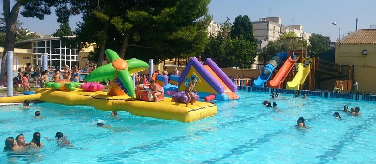 Муниципальный бассейн Parque del Oeste в Валенсии летом: яркие надувные горки в воде, дети плавают между ними, на заднем плане — деревья и крыши района L’Olivereta