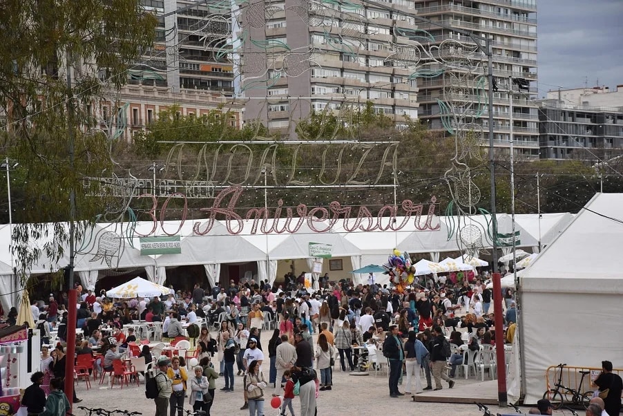 Feria Andaluza в Валенсии: вход с гирляндой огней «ANDALUZA Aniversari», ряды белых каcет с тапас, столики на улице, гуляющая публика, на заднем плане жилые дома района Algirós